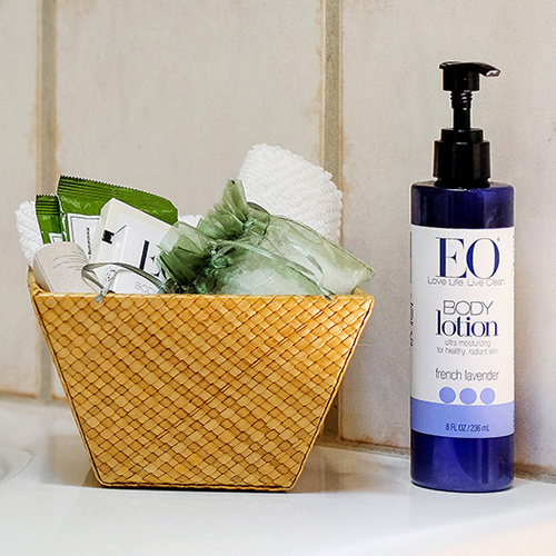 A basket with individually wrapped bath products and a bottle of EO Body Lotion set on the edge of the bathtubeo-bath-products-1up.jpg