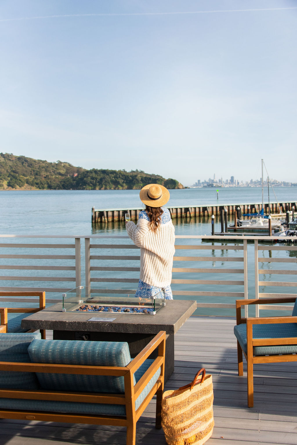 woman in flowy dress and sweater wearing a straw hat standing at the edge of the deck looking at the blue water below and city skyline in the distance on a sunny dayWEH-Deck-View-Kim.jpg