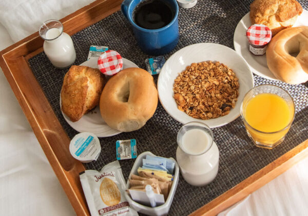 image of a wooden tray with breakfast small plates of food including bagels and pastries, oatmeal, small jars of jam, coffee, orange juice and milk pitcher. a person is perched next to the tray reading the newspaper.