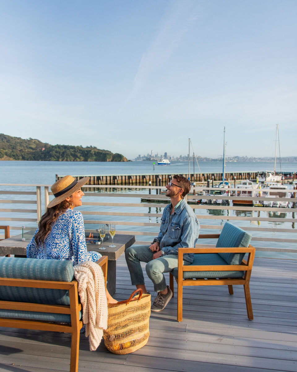 a man and a woman sitting on comfortable furniture on a deck overlooking the water and the city skyline in the distance. they are facing each other, laughing. there is a fire lit in the fire feature and there are glasses of champagne on the table.