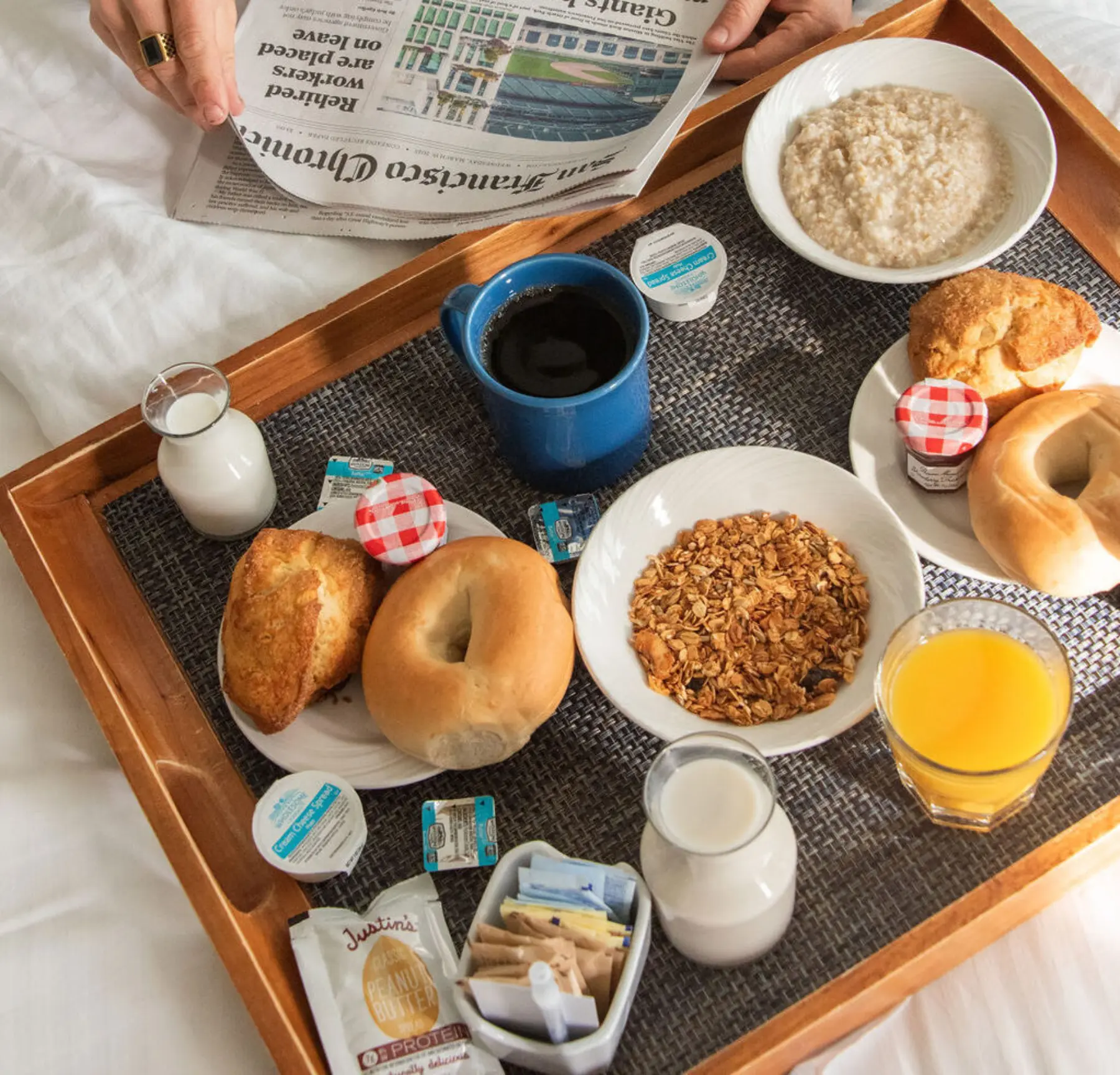 image of a wooden tray with breakfast small plates of food including bagels and pastries, oatmeal, small jars of jam, coffee, orange juice and milk pitcher. a person is perched next to the tray reading the newspaper.weh-breakfast-tray.jpg