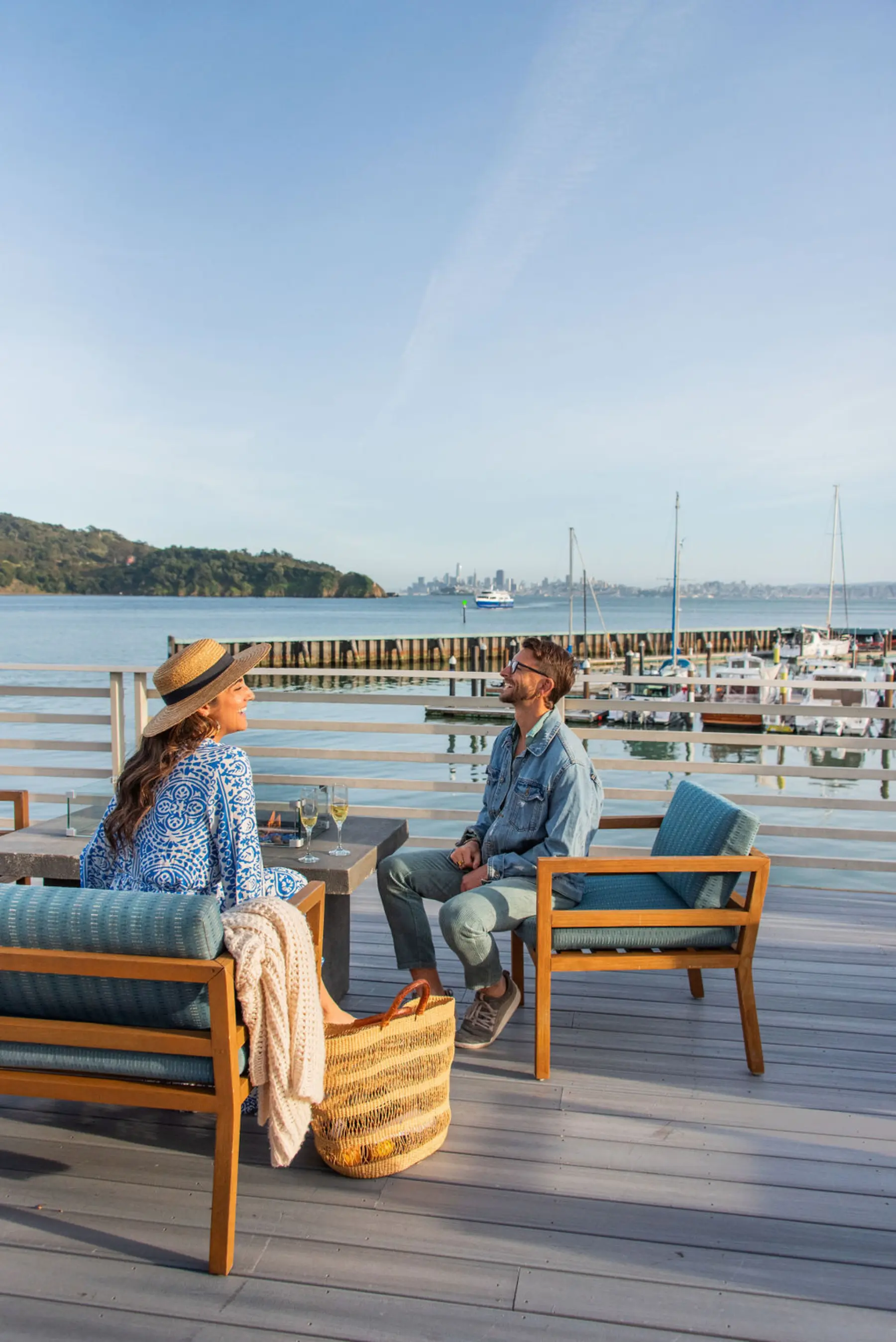 a man and a woman sitting on comfortable furniture on a deck overlooking the water and the city skyline in the distance. they are facing each other, laughing. there is a fire lit in the fire feature and there are glasses of champagne on the table.WEH-Kim-and-Nash-on-Deck.jpg